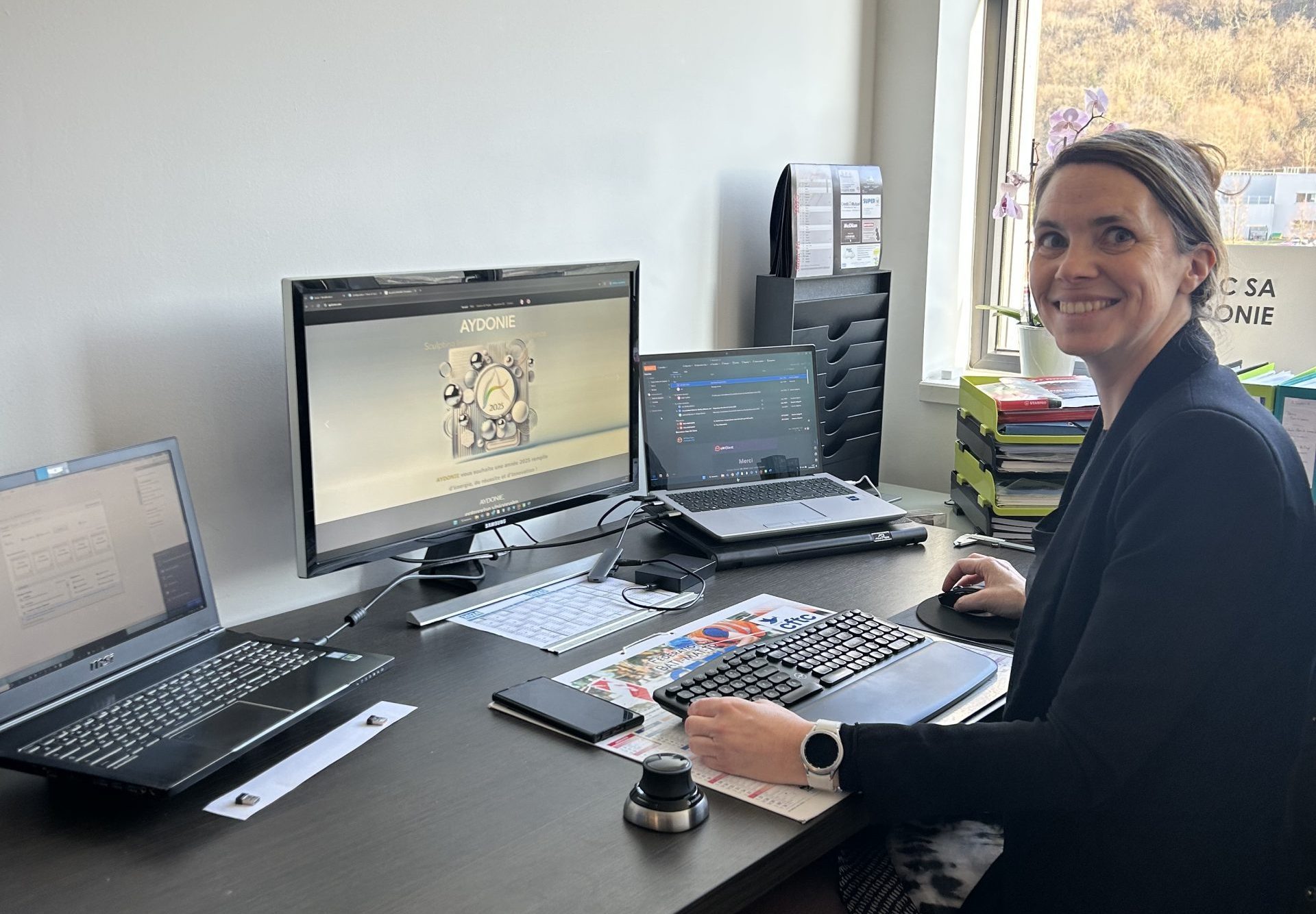 Femme blonde souriante assise à son bureau, devant un ordinateur.